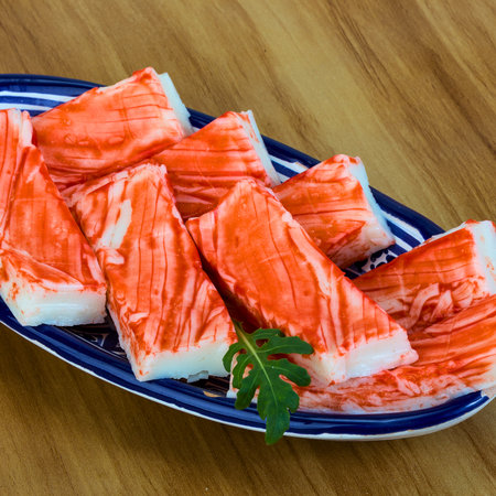 A close-up shot of bright red and white imitation crab sticks arranged on a decorative blue and white oval platter, garnished with a sprig of green herb.の写真素材