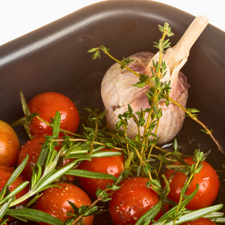 Close-up of ripe cherry tomatoes with fresh garlic and rosemary sprigs in a dark pan, ready for cooking.の写真素材