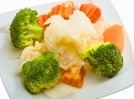 A close-up shot of a healthy mix of steamed broccoli florets, carrot chunks, and cauliflower pieces arranged on a white plate.の写真素材