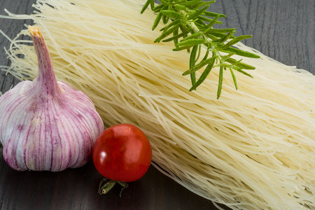 A close-up shot of raw ingredients including a garlic bulb, a cherry tomato, and a bundle of thin noodles, with a sprig of rosemary.の写真素材