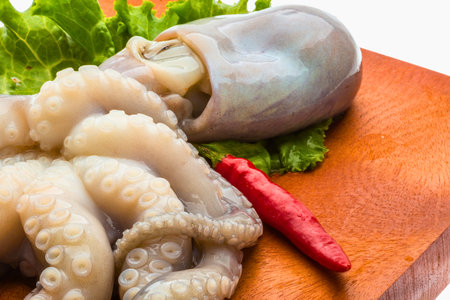 A close-up shot of a raw octopus with its tentacles spread out, accompanied by a red chili pepper and greens on a wooden cutting board.の写真素材