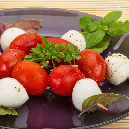 A vibrant Caprese salad featuring ripe cherry tomatoes, creamy mozzarella balls, and fresh basil leaves on a dark plate.の写真素材