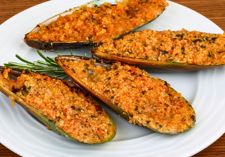 Four savory baked mussels are generously stuffed with a flavorful mixture and garnished with fresh rosemary on a white plate.の写真素材