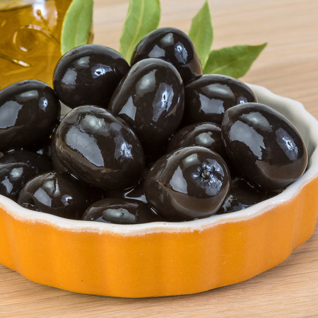 A close-up shot of plump, glossy black olives piled high in a decorative orange bowl, with a hint of olive oil and green leaves in the background.の写真素材
