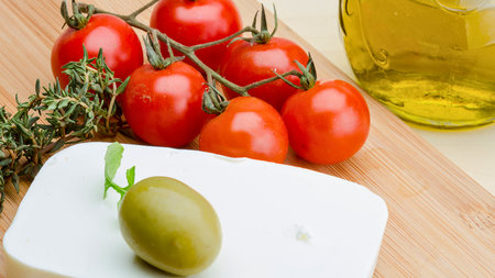 A still life of ripe cherry tomatoes on the vine, a single green olive, and a sprig of herbs next to a bottle of olive oil.の写真素材