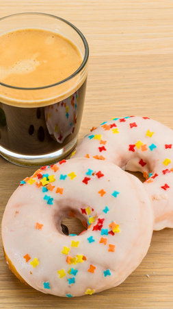 A close-up shot of two pink frosted donuts adorned with colorful sprinkles, placed next to a steaming cup of dark coffee.の写真素材