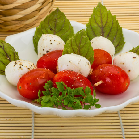 Appetizing skewers featuring fresh mozzarella balls, cherry tomatoes, and vibrant green basil leaves, artfully arranged on a white plate.の写真素材