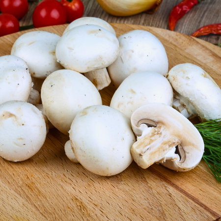 A pile of fresh white button mushrooms sits on a rustic wooden cutting board, with hints of red peppers and herbs in the background.の写真素材