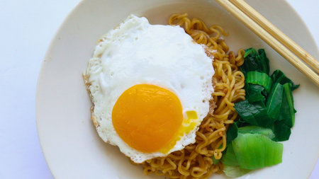 A close-up shot of a plate of instant noodles, a perfectly fried egg, and vibrant green vegetables,...
