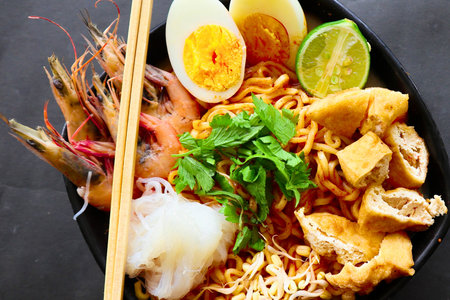 A close-up shot of a steaming bowl of spicy shrimp noodle soup, garnished with halved boiled eggs, lime wedges, and fresh herbs.の写真素材