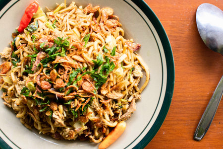 A close-up overhead view of a bowl filled with noodles, garnished with fresh herbs, chili, and fried shallots, next to a spoon.の写真素材
