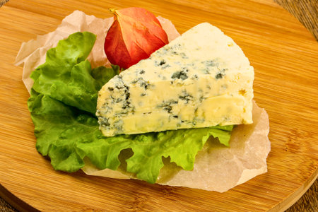 A wedge of blue cheese sits atop fresh green lettuce leaves, garnished with a single red fruit, presented on a wooden board.の写真素材