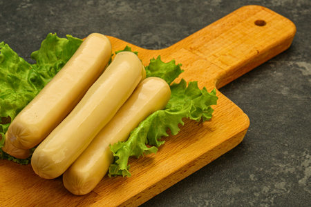 Three light-colored, boiled sausages arranged on fresh green lettuce, served on a natural wooden cutting board. Ideal for food advertising or healthy meal concepts.の写真素材
