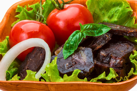 A rustic wooden bowl holds savory cured meat slices, vibrant red tomatoes, crisp green lettuce, a white onion ring, and a fragrant basil leaf.の写真素材