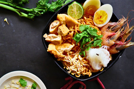 Top-down view of a vibrant bowl of spicy Asian noodles featuring fresh shrimp, crispy tofu, hard-boiled eggs, bean sprouts, and a refreshing lime wedge.の写真素材