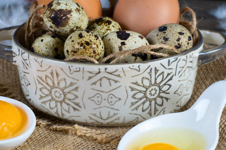 Assortment of fresh eggs, including speckled quail eggs in a decorative bowl, brown chicken eggs, and raw egg yolk and white in small dishes, on burlap.の写真素材