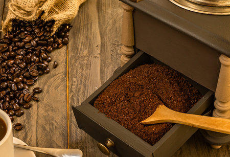Dark roasted coffee beans from burlap sack on rustic wood. Antique grinder, fresh grounds, scoop. Coffee cup evokes fresh morning aroma, homemade brew.の写真素材