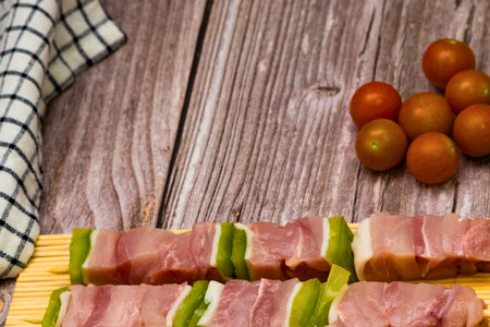 Freshly prepared raw meat and green bell pepper skewers arranged on a wooden board, accompanied by vibrant red cherry tomatoes, ready for grilling.の写真素材