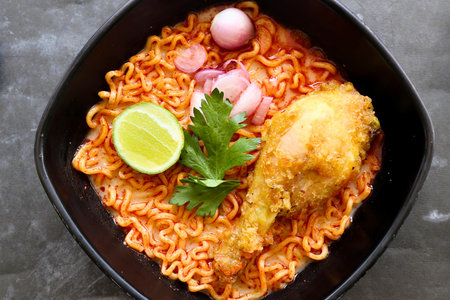 Top view of spicy chicken ramen noodles with crispy fried drumstick, lime, and shallots in a black bowl. A savory and comforting Asian dish, rich in flavor, ready to enjoy.の写真素材
