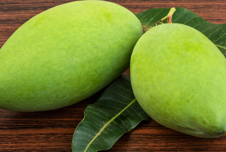 A close-up shot of two fresh, vibrant green mangoes accompanied by their leaves, resting on a rustic dark wooden surface. These tropical fruits are popular globally.の写真素材