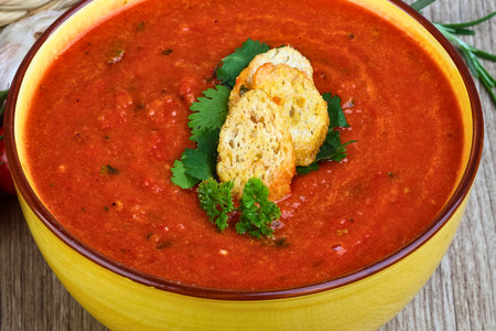 Rich red tomato soup in a vibrant yellow bowl, adorned with crispy golden croutons and fresh green herbs. A healthy, warm, and comforting meal on a wooden surface, perfect for cozy dining.の写真素材