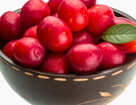 A close-up shot of a dark bowl filled with bright red, oval-shaped cornelian cherries, one green leaf visible.の写真素材