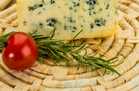 A close-up shot of a creamy blue cheese wedge, a bright red cherry tomato, and a sprig of fresh rosemary resting on a woven surface.の写真素材