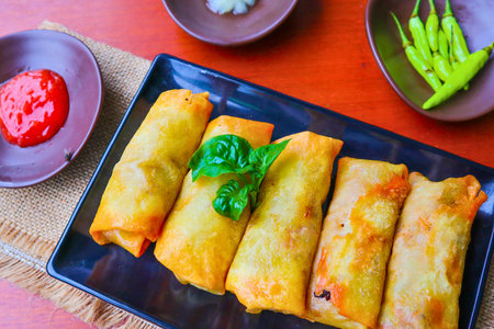 A close-up overhead shot of golden fried spring rolls on a black rectangular plate, served with a red dipping sauce and a green garnish.の写真素材