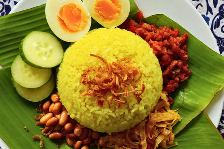 A colorful and appetizing plate of yellow rice, hard-boiled eggs, sliced cucumbers, peanuts, and shredded chicken served on banana leaves.の写真素材