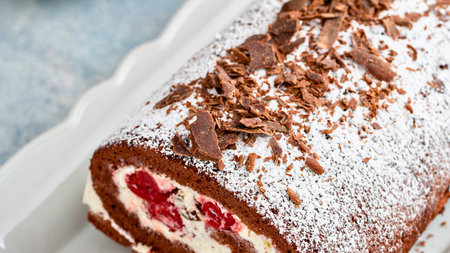 A close-up view of a chocolate Swiss roll cake filled with whipped cream and cherries, dusted with powdered sugar and topped with chocolate shavings.の写真素材