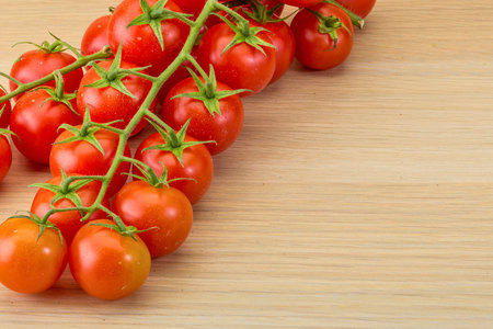 A cluster of ripe, red cherry tomatoes hangs on a vine against a light wooden surface, ready for use in cooking.の写真素材