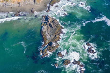 An overhead drone shot captures a rugged rock formation surrounded by crashing turquoise waves and white sea foam along a sandy coastline.の写真素材