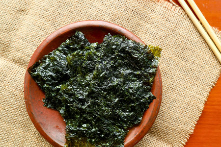 A close-up overhead view of a pile of dark green, crispy roasted seaweed presented in a reddish-brown ceramic dish on a textured burlap surface.の写真素材