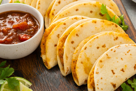 A close-up view of several golden brown crispy tacos arranged on a wooden board next to a bowl of chunky salsa and lime wedges.の写真素材