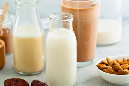 Several glass bottles and a glass filled with various plant based milks, alongside almonds and dates.の写真素材