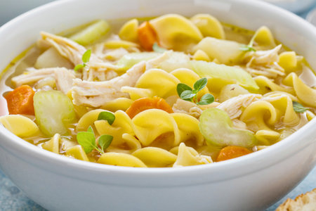 A close-up shot of a steaming bowl of chicken noodle soup, packed with shredded chicken, pasta, carrots, and celery.の写真素材