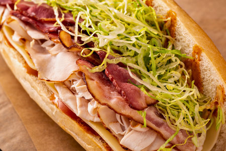 A close-up view of a long sub sandwich filled with layers of deli meats, bacon, and shredded lettuce on a golden bun.の写真素材