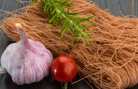 A close-up shot of uncooked pasta, a head of garlic, a tomato, and a sprig of rosemary on a dark wooden background.の写真素材