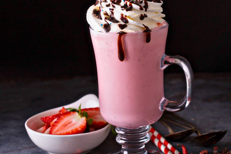 A tall glass of creamy pink strawberry milkshake topped with whipped cream and chocolate syrup, served with fresh strawberries.の写真素材