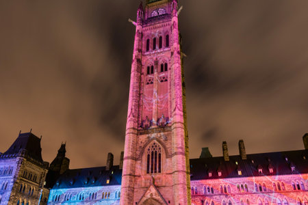 The iconic Peace Tower of the Canadian Parliament Building is dramatically lit with vibrant pink and blue lights against a cloudy night sky.の写真素材
