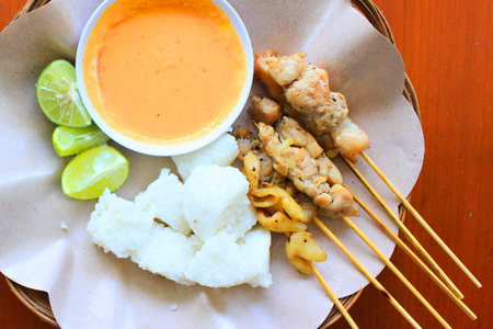 A close-up overhead view of grilled chicken satay skewers served with a creamy peanut sauce, lime wedges, and a side of rice.の写真素材