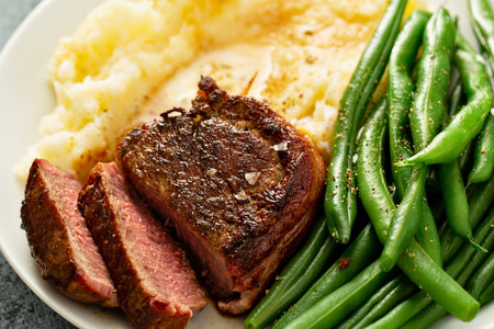 A close-up shot of a perfectly cooked sliced steak served with creamy mashed potatoes and fresh green beans on a white plate.の写真素材