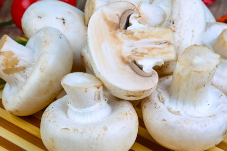 A close-up shot of several fresh, whole white button mushrooms, with one cut in half revealing its gills. The mushrooms are piled on a wooden surface.の写真素材