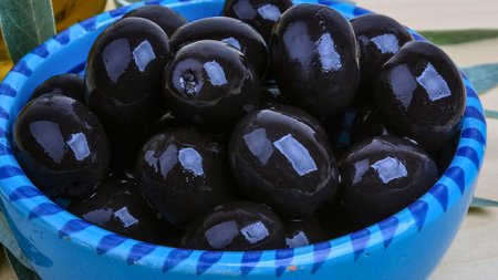 A vibrant close-up showcases a collection of glossy, dark black olives nestled within a bright blue and white checkered bowl.の写真素材