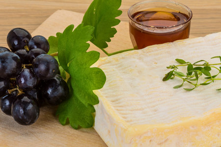 A close-up of a creamy cheese wedge with fresh grapes, a small glass of amber liquid, and green herbs on a wooden surface.の写真素材
