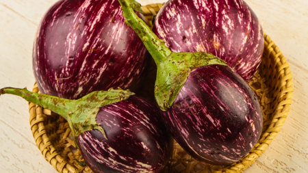 A close-up overhead view of several ripe, striped graffiti eggplants nestled in a rustic woven basket on a textured surface.の写真素材