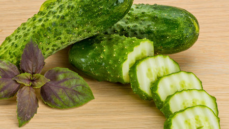A close-up shot of whole and sliced cucumbers alongside a sprig of purple basil on a light-colored wooden background.の写真素材
