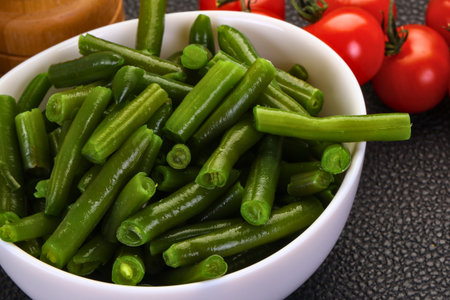 A close-up shot of vibrant green beans, lightly cooked and glistening, served in a white bowl. Ripe red tomatoes are visible in the background.の写真素材