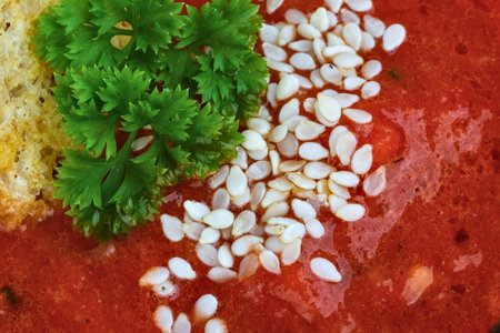 A textured, close-up view of a rich red soup, artfully decorated with bright green parsley and scattered white seeds.の写真素材