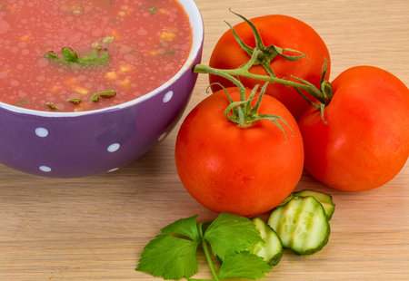 A bowl of vibrant red gazpacho soup sits beside ripe tomatoes, sliced cucumber, and fresh basil leaves on a wooden surface.の写真素材